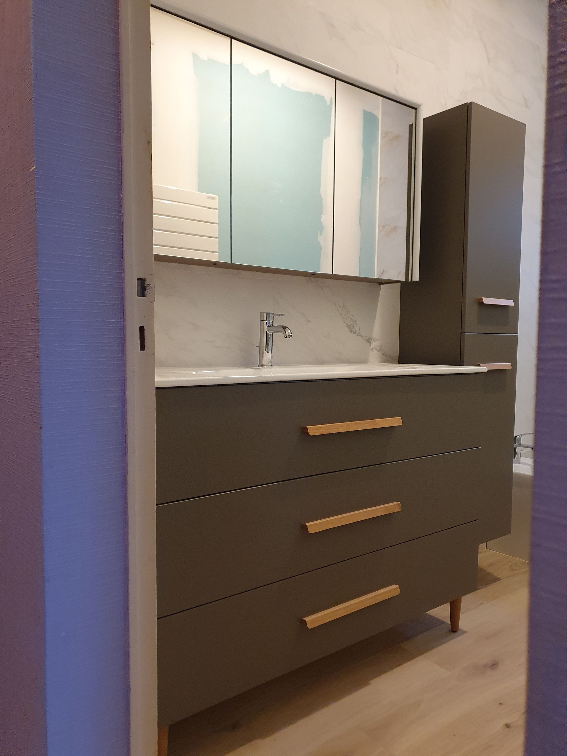 Meuble-lavabo moderne avec tiroirs gris, poignées en bois clair, miroir et armoire haute.