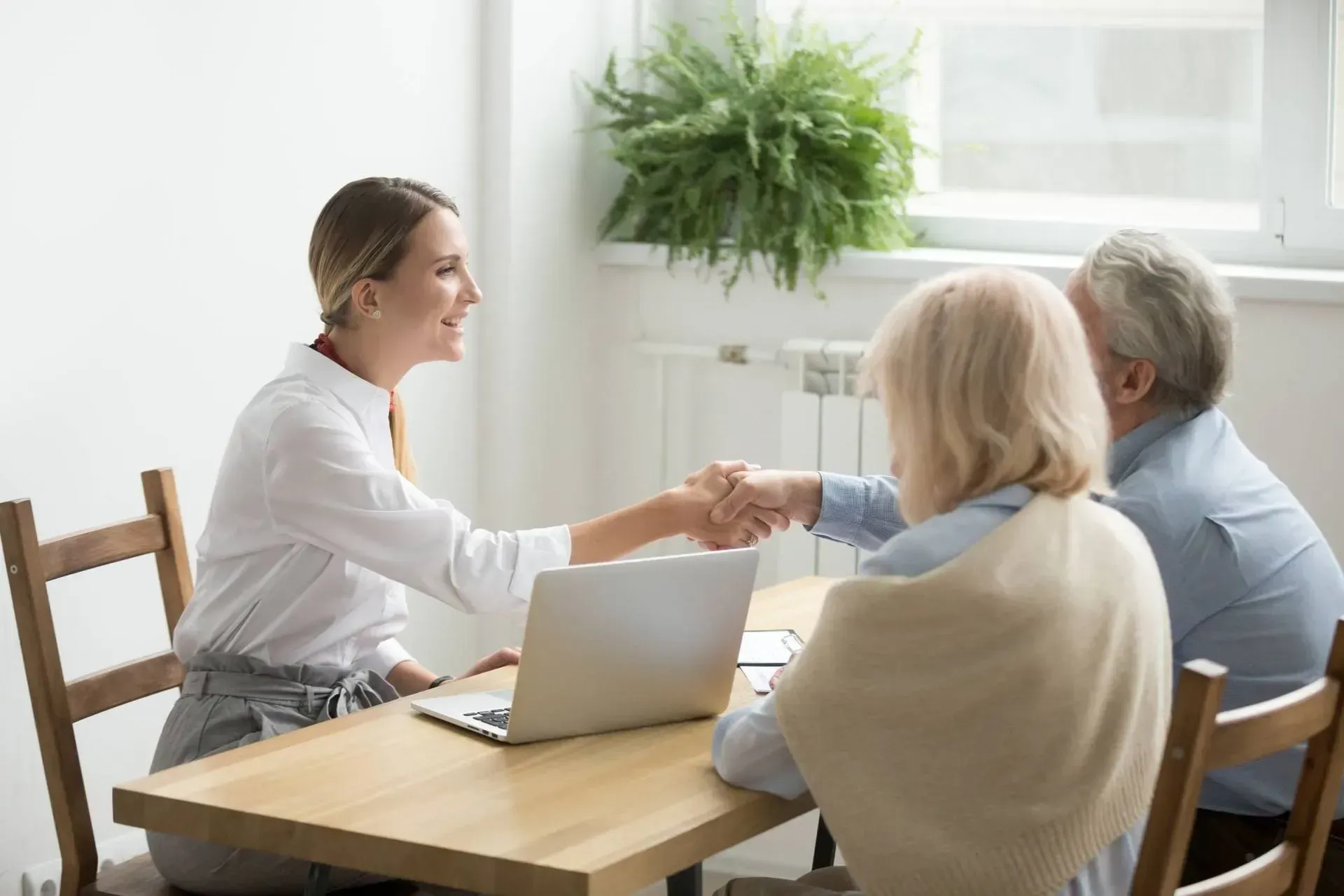 Une jeune femme serre la main d'une personne âgée à une table. Ordinateur portable, papiers, sourires et fenêtre.