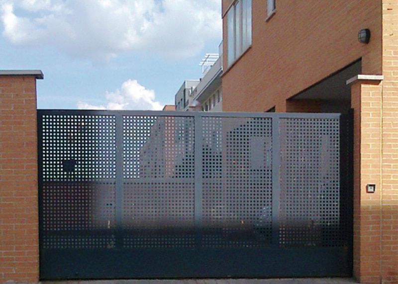Puerta de seguridad metálica con patrón de rejilla, situada entre pilares de ladrillo, frente a un edificio.