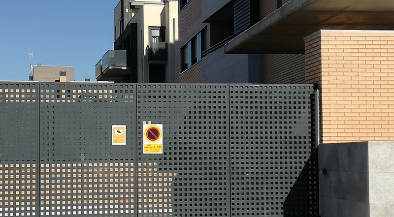 Puerta metálica gris con recortes cuadrados y señales de prohibido aparcar. Edificio de ladrillo y moderno al fondo.