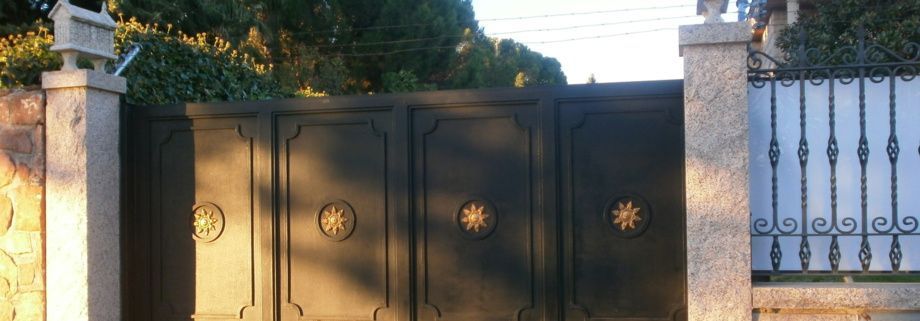Una puerta de metal oscuro con adornos decorativos en forma de estrella, flanqueada por pilares de piedra y vegetación.