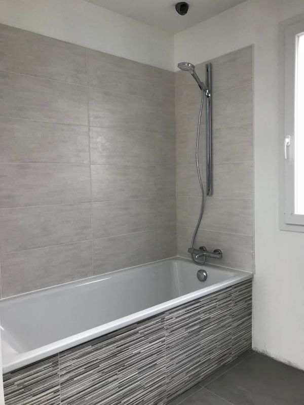 Salle de bains avec murs carrelés gris, baignoire blanche, pommeau de douche et fenêtre.