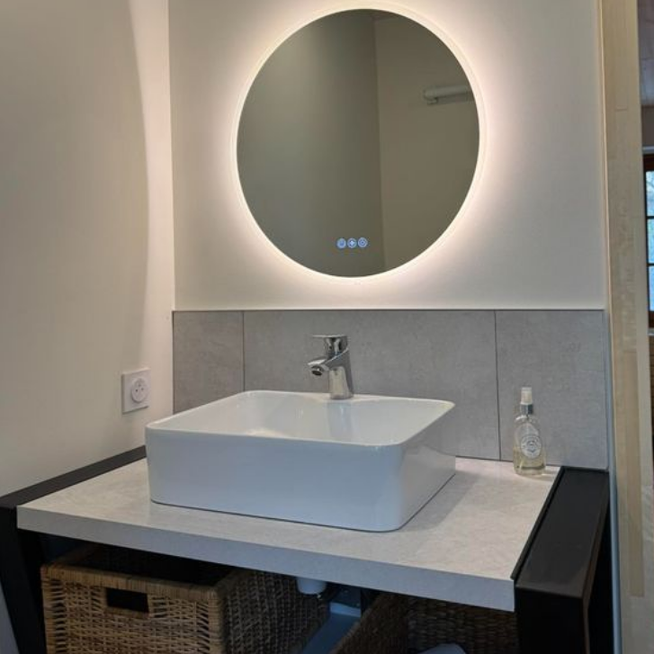 Lavabo de salle de bain avec vasque carrée, robinet moderne et miroir rond à bord lumineux.