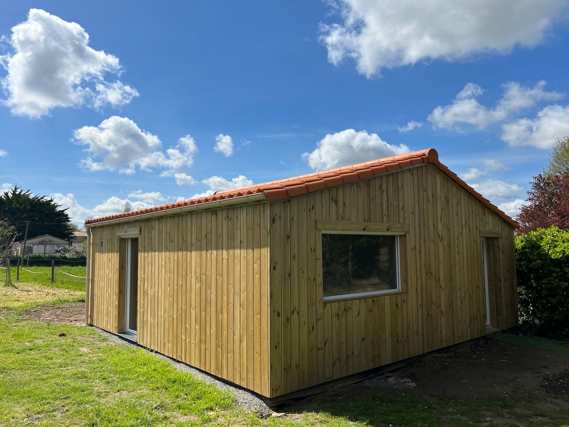 Cabane en bois au toit de tuiles orange et deux fenêtres sous un ciel bleu parsemé de nuages ​​blancs duveteux.