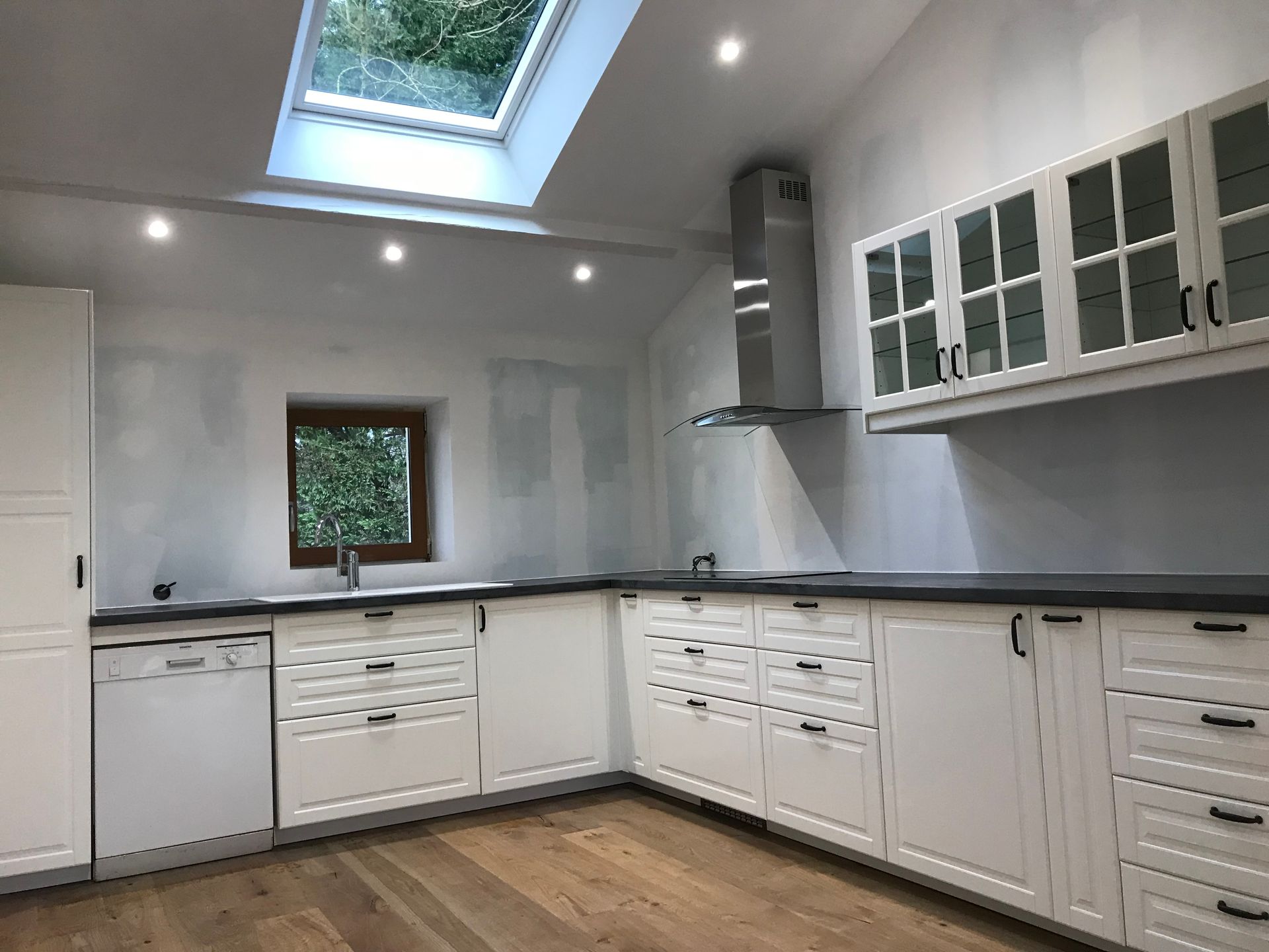 Cuisine blanche avec comptoirs noirs, plancher en bois, fenêtre et puits de lumière.