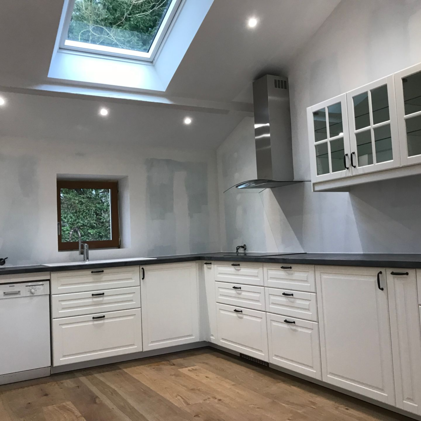 Cuisine blanche avec puits de lumière, placards et électroménager. Sol en bois et fenêtre donnant sur la verdure.