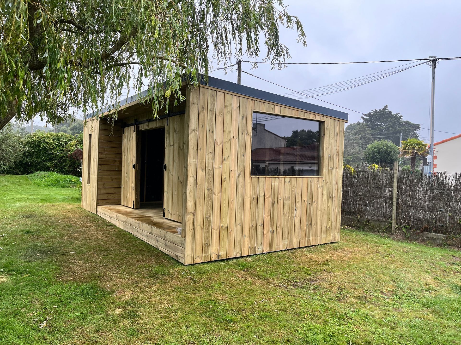 Abri de jardin en bois avec un toit sombre et une fenêtre, dans une cour herbeuse sous un arbre.
