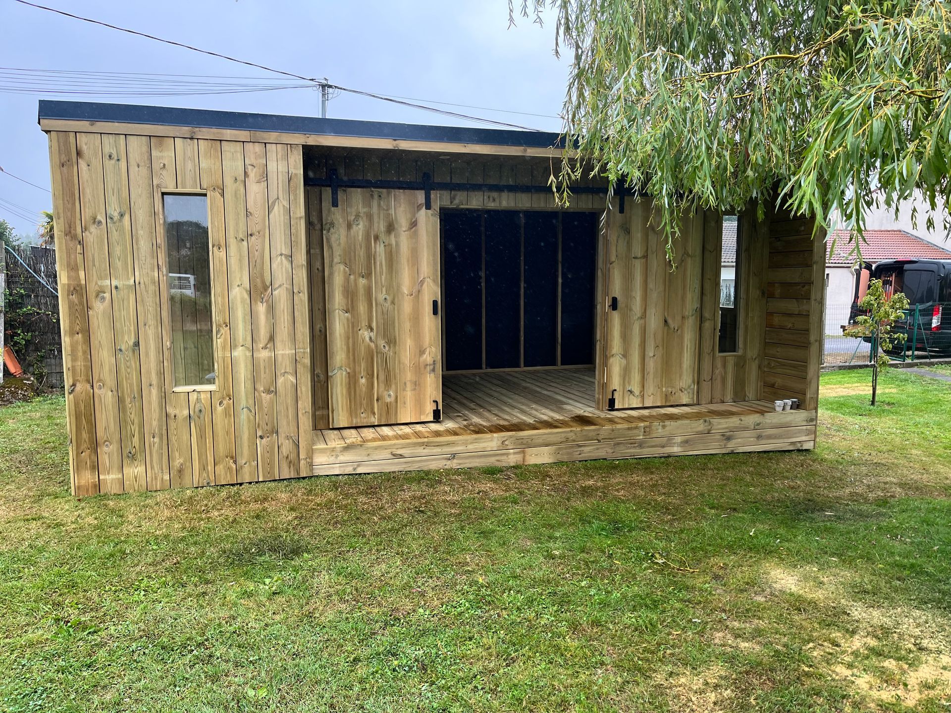 Un abri en bois avec portes coulissantes et deux fenêtres se trouve sur l'herbe verte à côté d'un arbre.