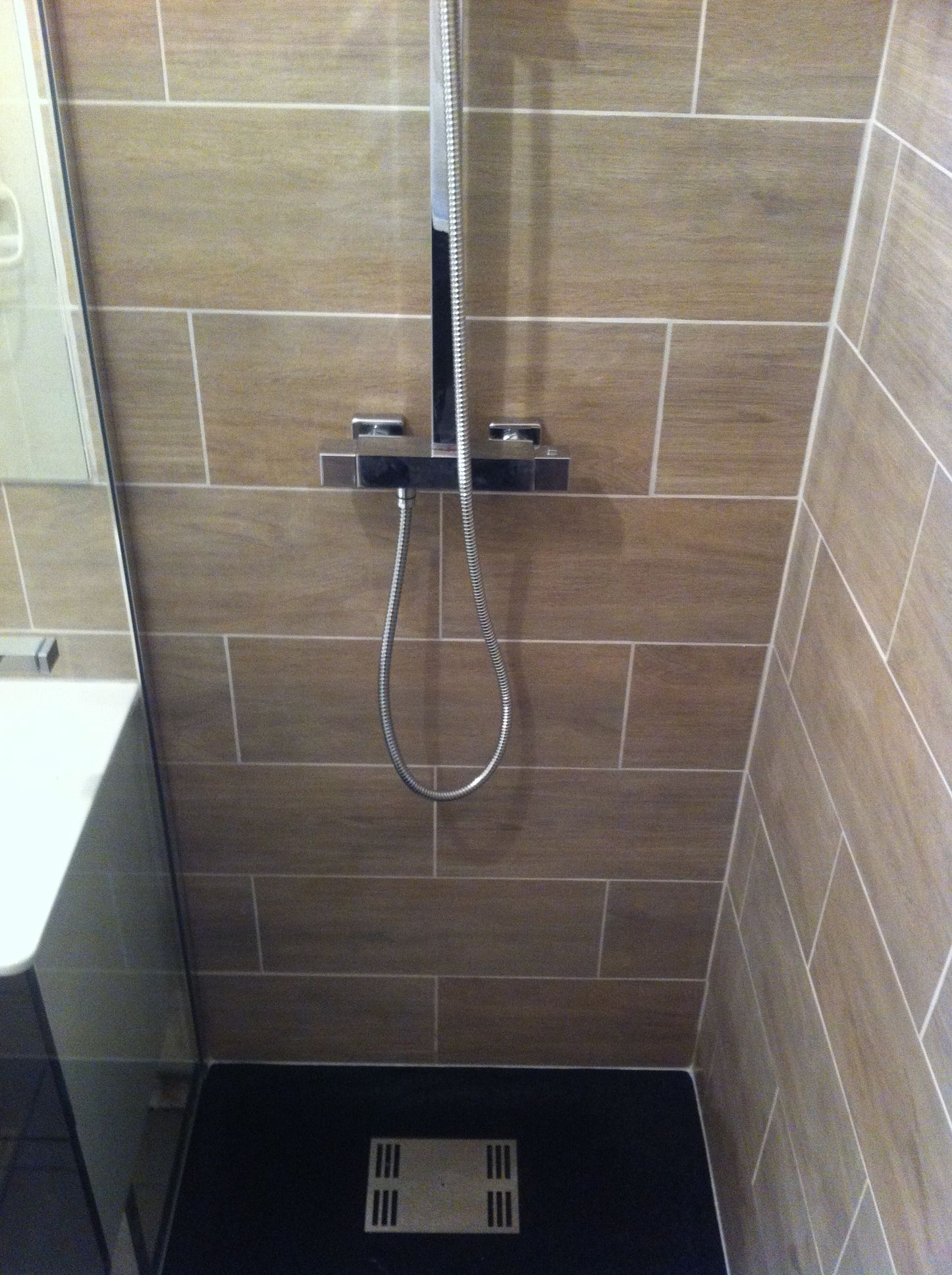 Cabine de douche avec murs carrelés marron et sol foncé. Pommeau et poignée de douche chromés.