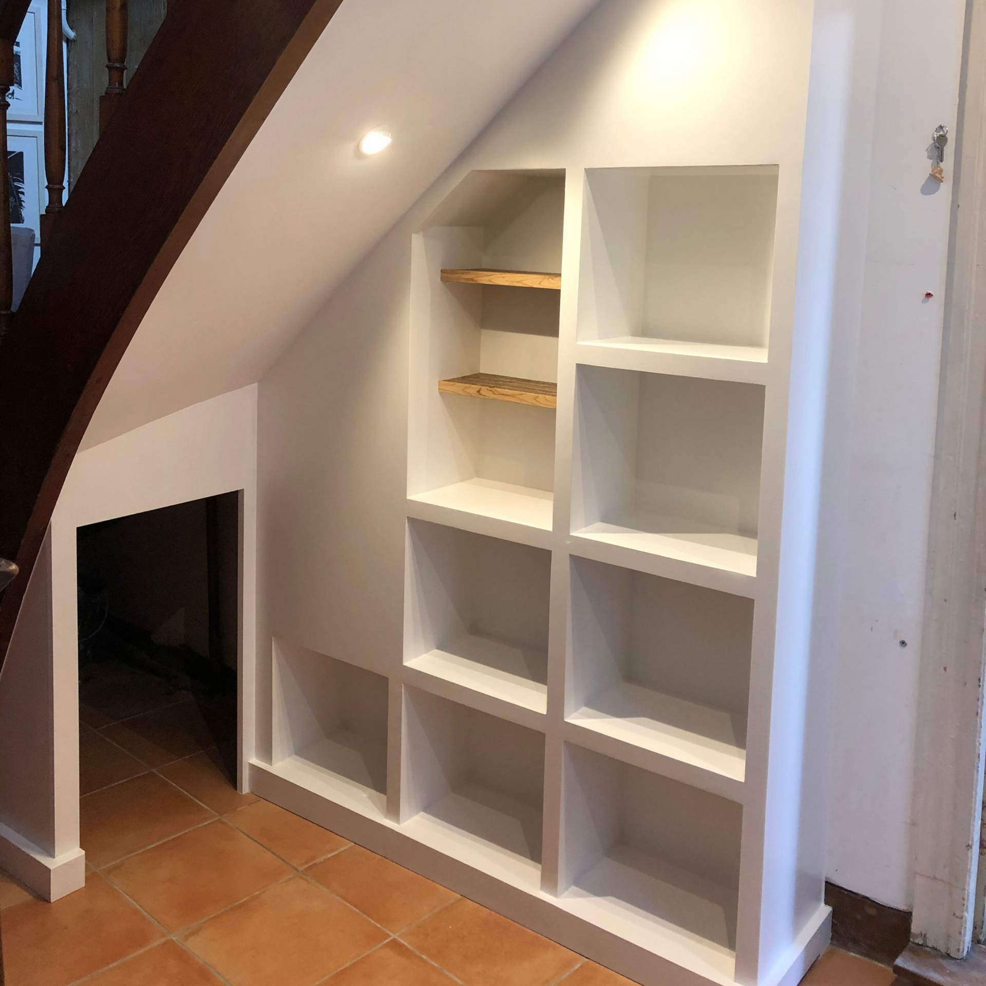 Étagères blanches encastrées sous l'escalier, avec une ouverture. On aperçoit un sol carrelé marron, des murs blancs et un escalier en bois foncé.