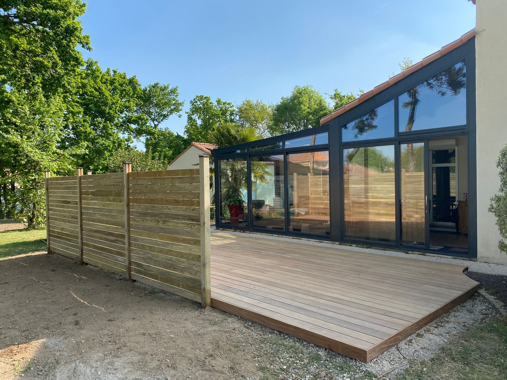 Terrasse en bois et clôture longeant une véranda vitrée par une journée ensoleillée.