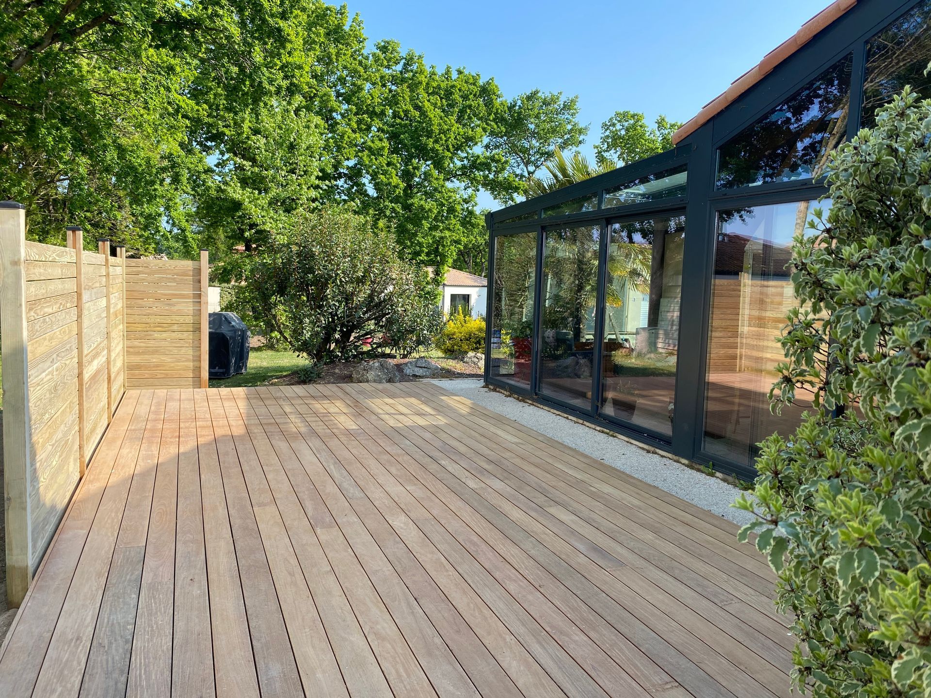 Terrasse en bois jouxtant une véranda vitrée et une clôture en bois, sous un ciel bleu.