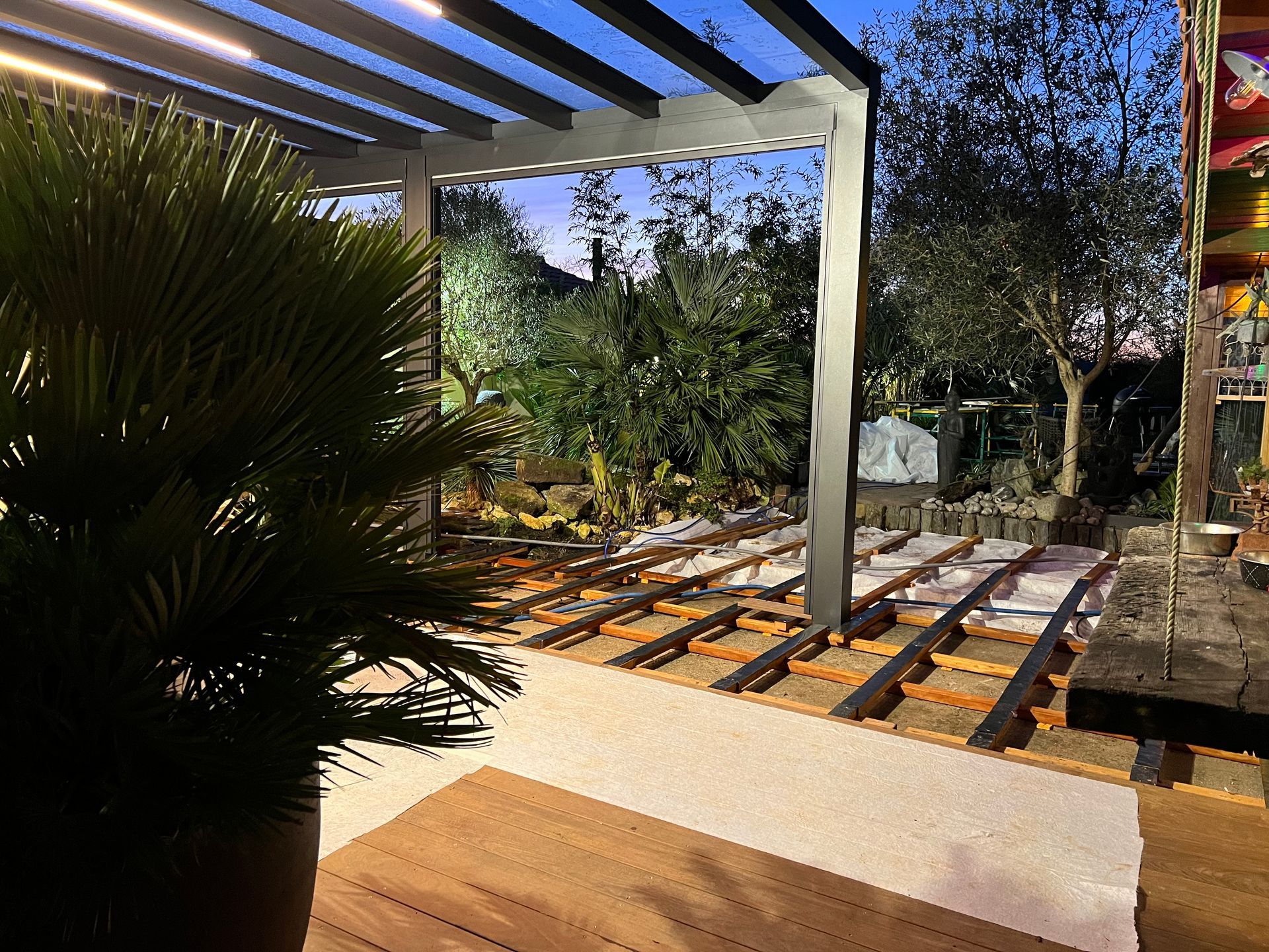 Pergola extérieure avec terrasse apparente, palmier au premier plan, ambiance crépusculaire.