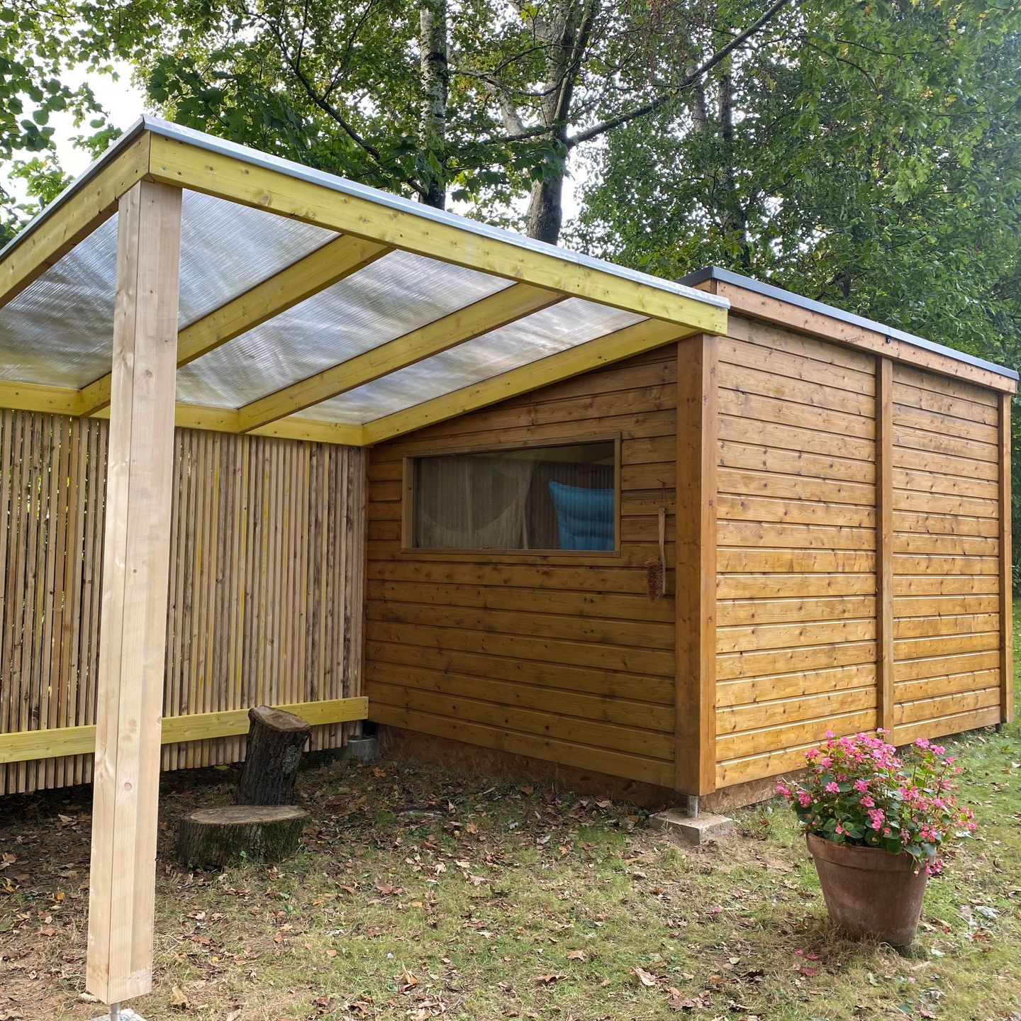 Abri de jardin en bois avec une entrée couverte et un pot de fleurs devant.