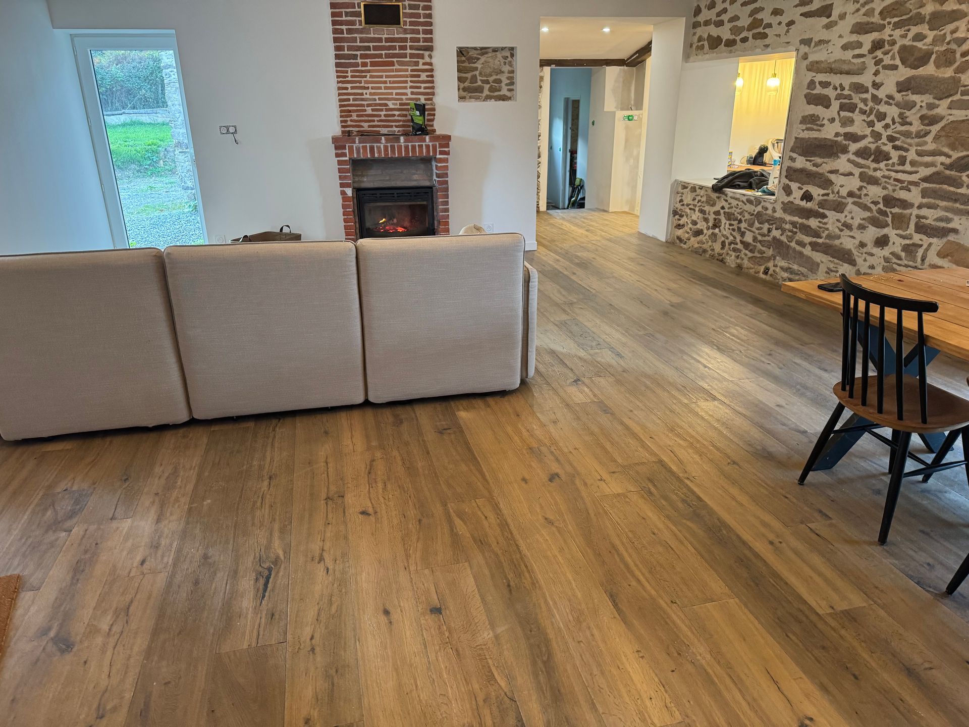 Salon avec parquet clair, canapé beige, cheminée en briques et mur en pierre.