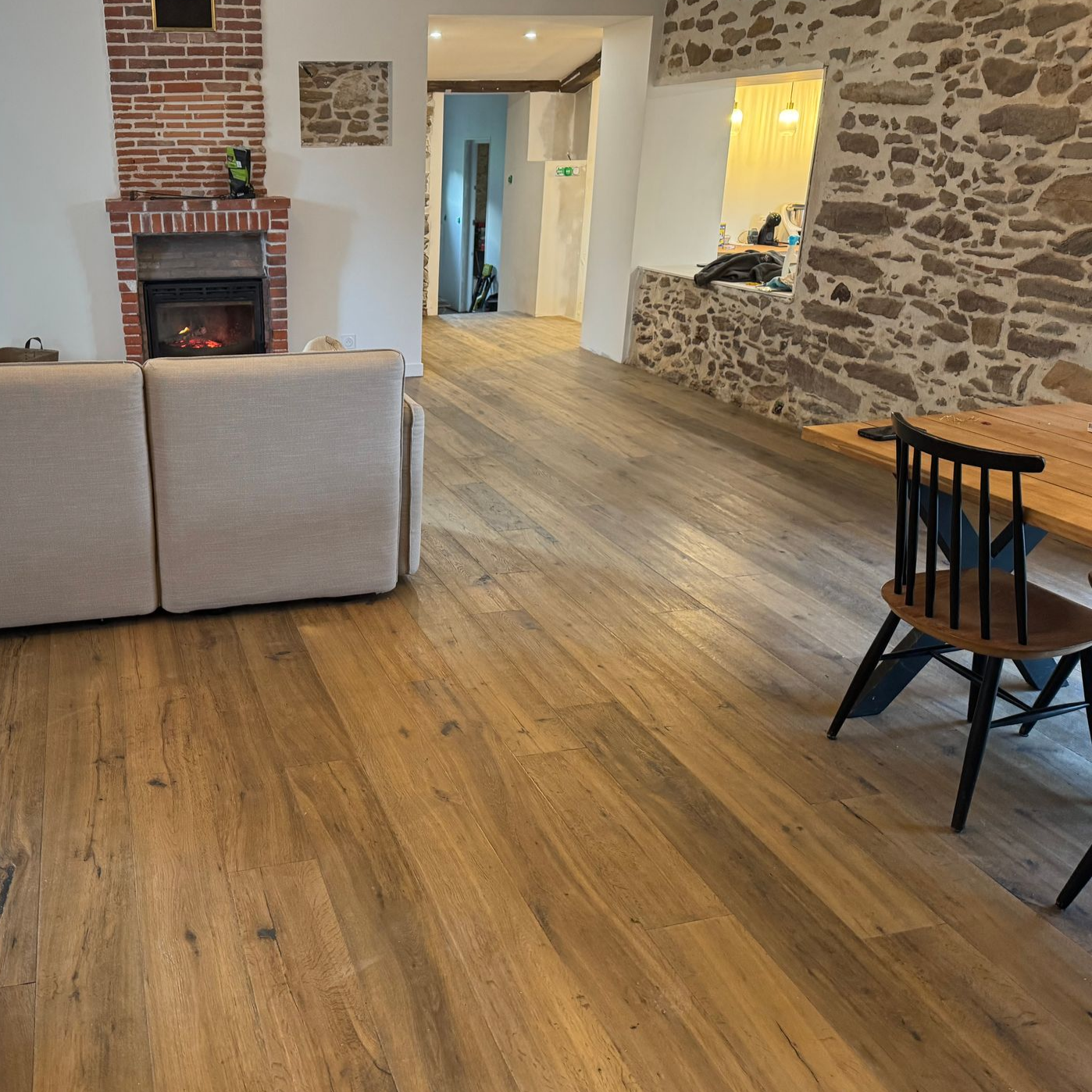 Pièce spacieuse avec parquet, cheminée et mur en pierre apparente. Table à manger avec chaises noires.