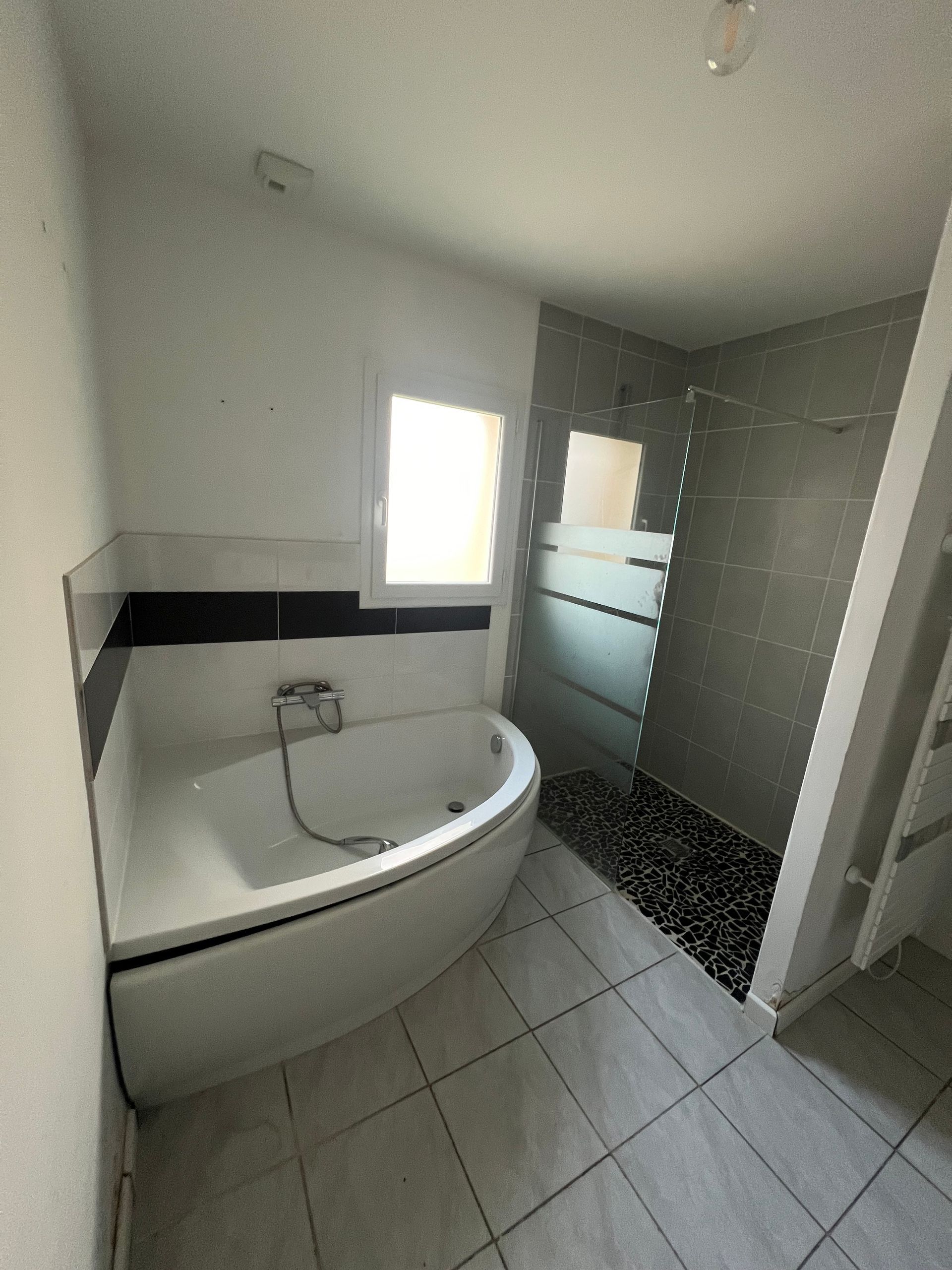 Salle de bains avec baignoire blanche, douche en verre et sol carrelé gris. Une fenêtre apporte de la lumière naturelle.