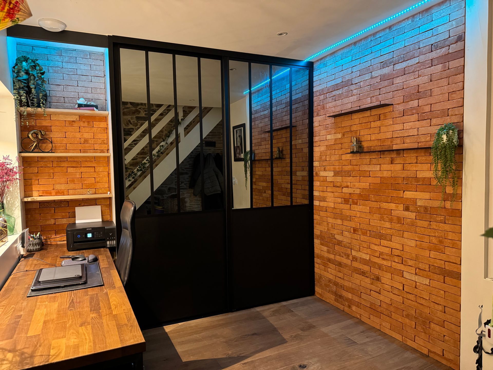 Bureau à domicile avec murs en briques apparentes, porte vitrée à cadre noir, bureau en bois et éclairage LED bleu.