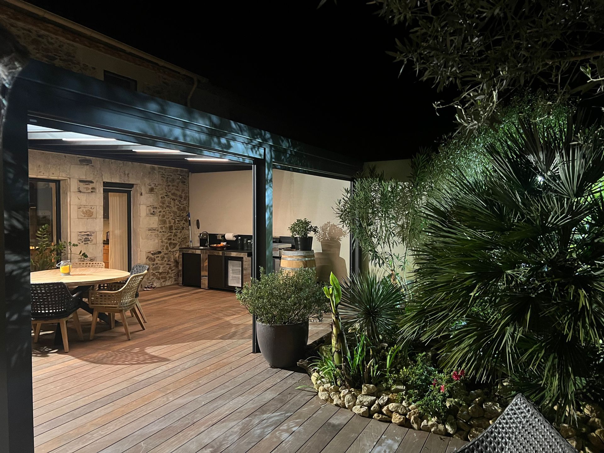 Terrasse nocturne avec plancher en bois, table à manger, espace barbecue et végétation luxuriante.