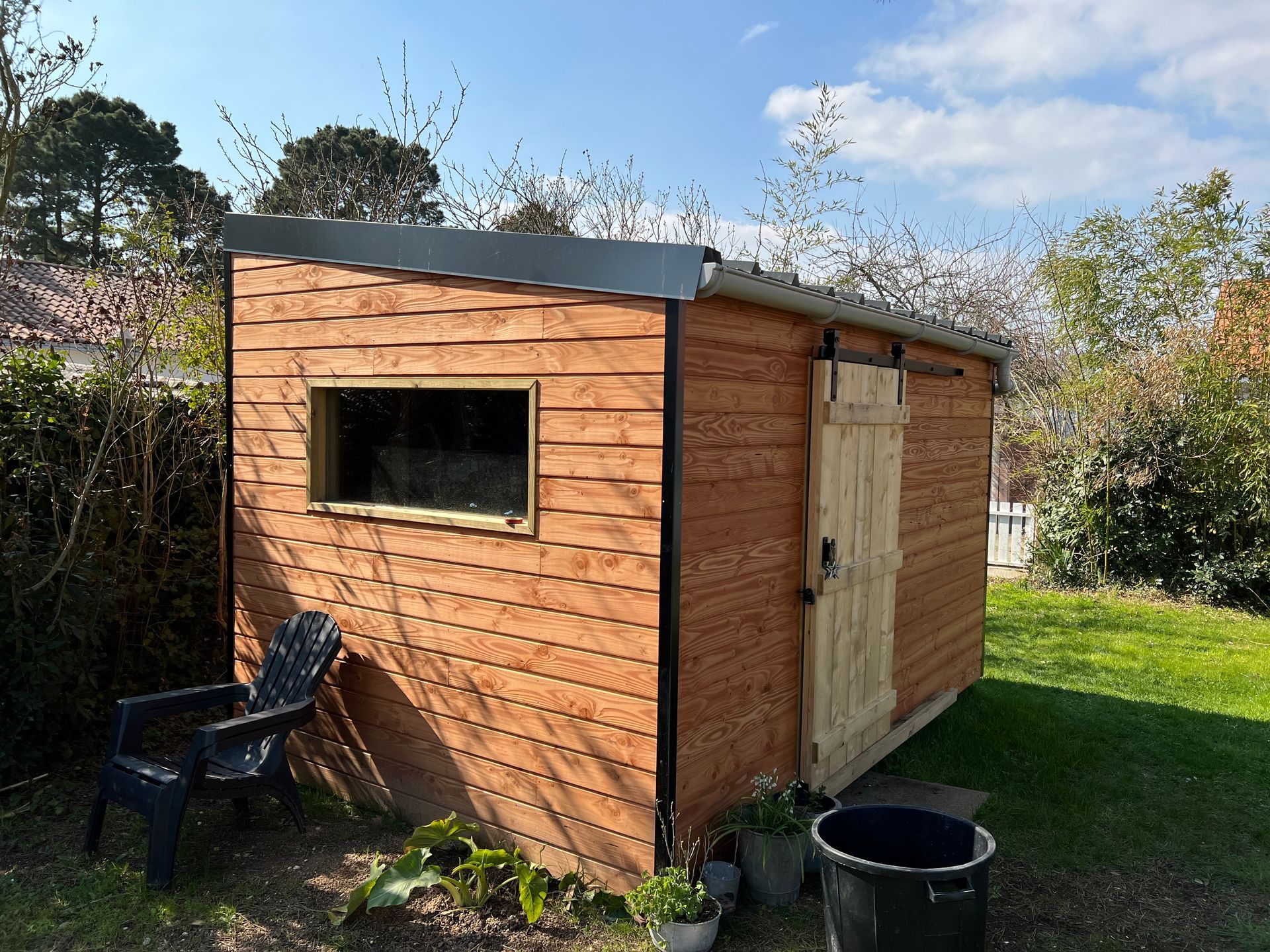 Abri de jardin en bois avec porte coulissante, fenêtre et chaise noire dans une cour herbeuse.