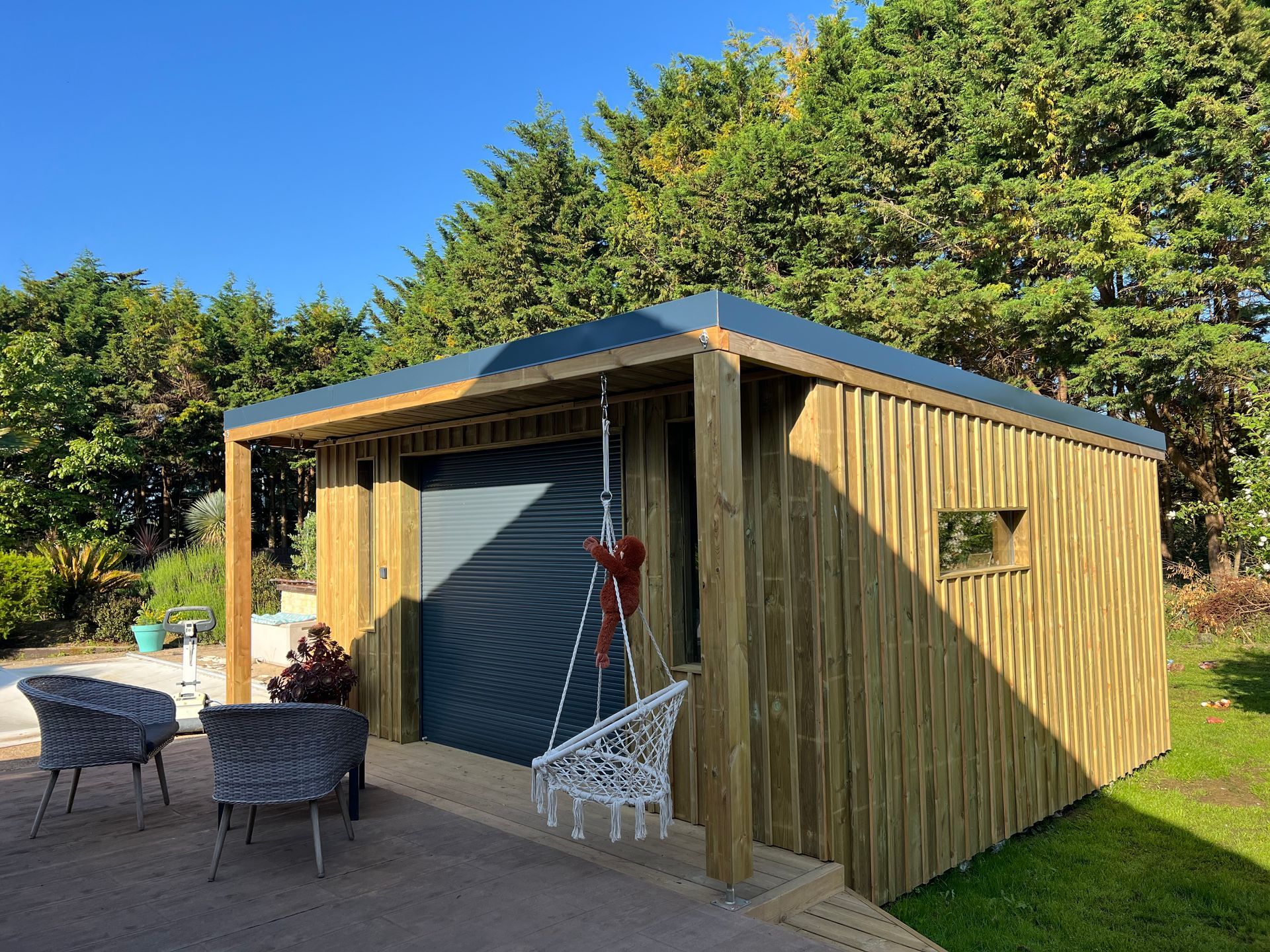 Abri de jardin en bois avec toit gris, porche ouvert, balançoire et chaises en osier.