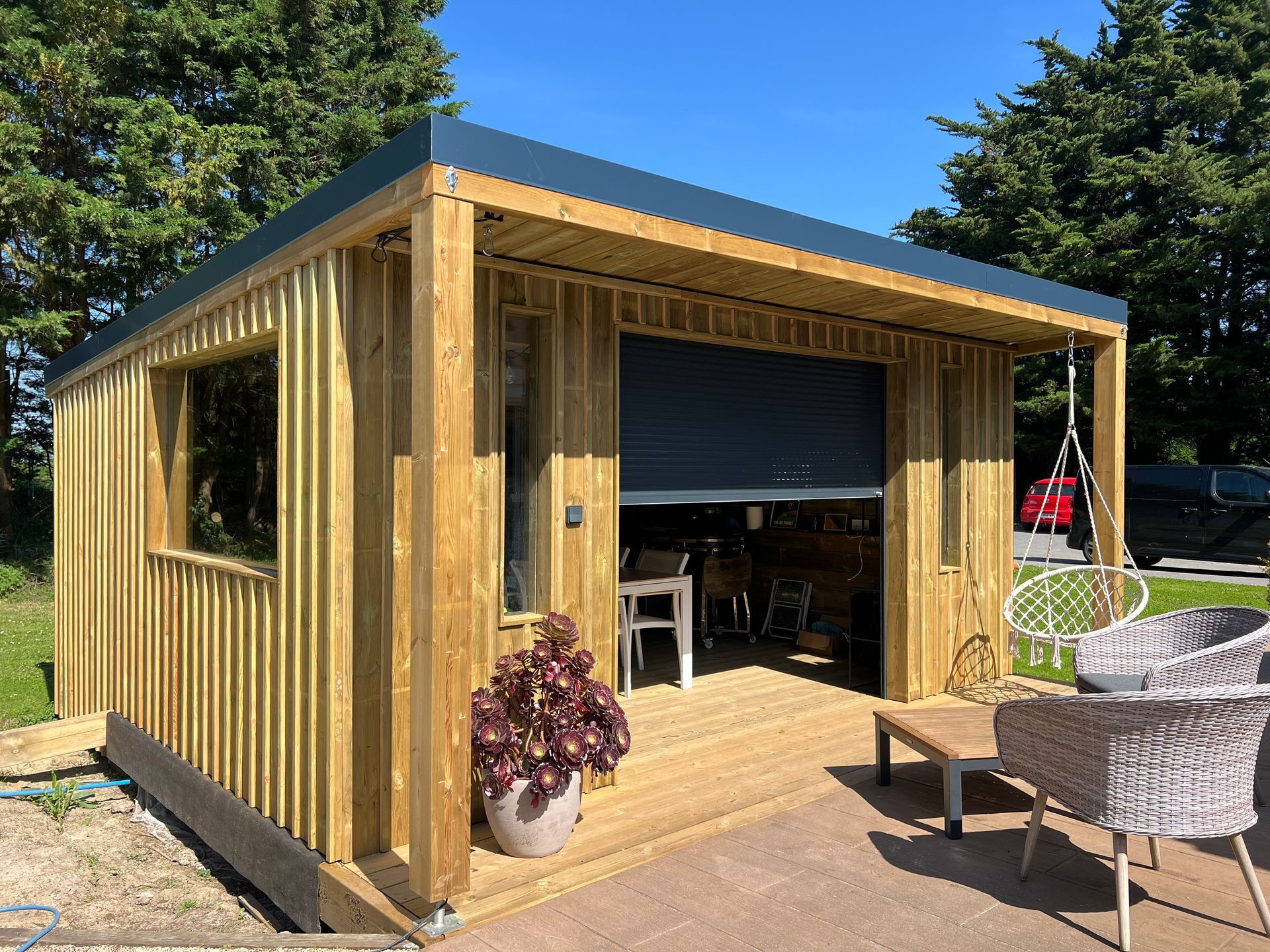 Cabane en bois au toit gris foncé et volet partiellement ouvert. Extérieur ensoleillé.