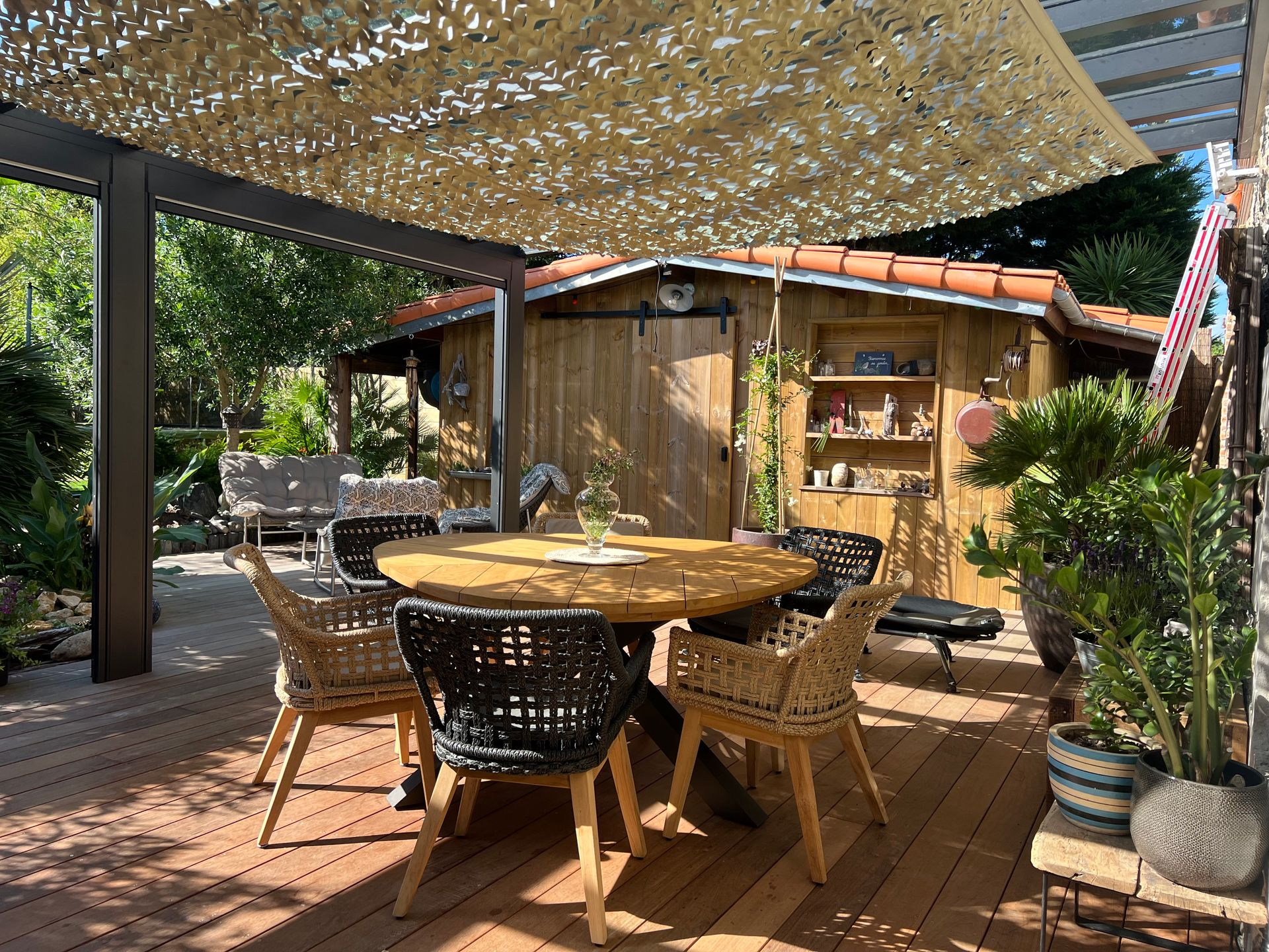 Terrasse avec table ronde, chaises et auvent près d'un abri en bois.