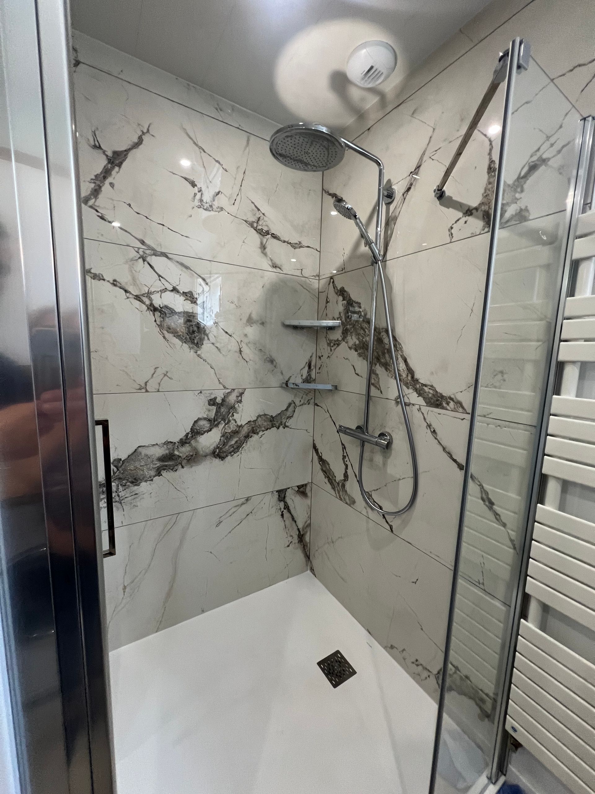 Cabine de douche avec carrelage imitation marbre blanc et gris, porte en verre et robinetterie chromée.