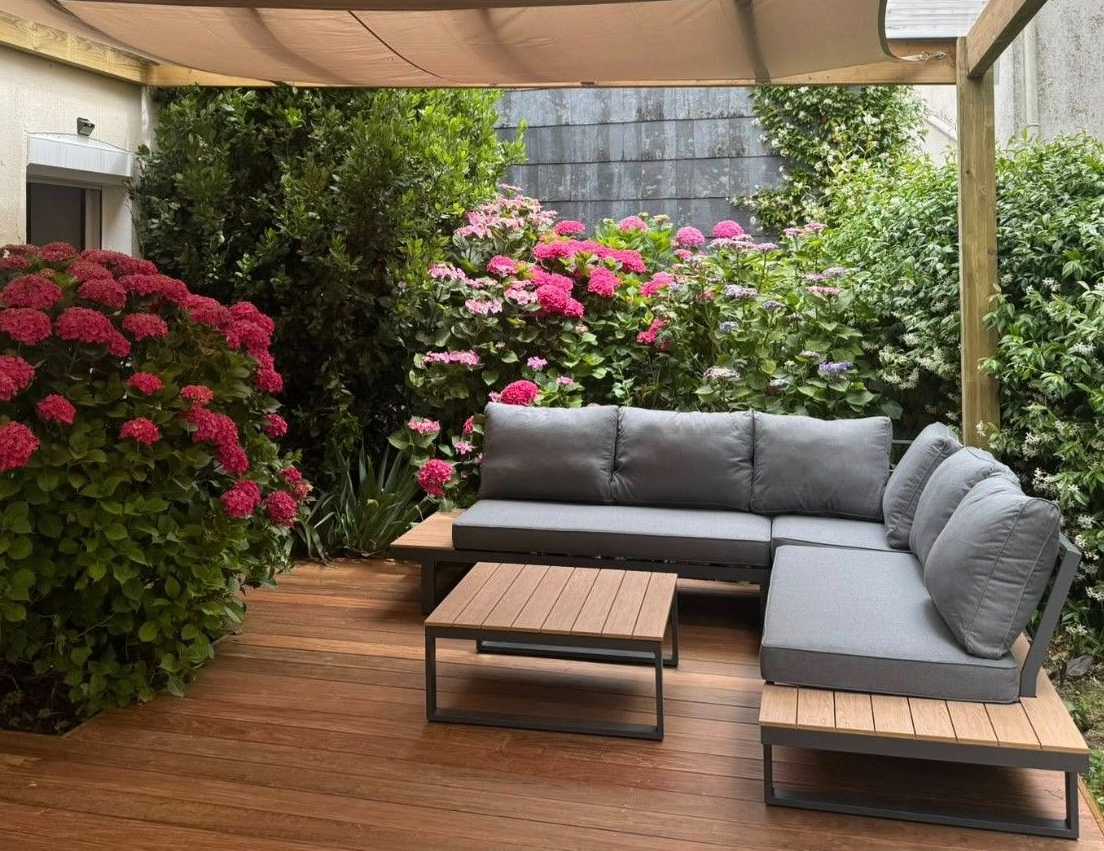 Espace détente extérieur avec canapé d'angle gris sur une terrasse en bois, entouré de fleurs et de verdure luxuriantes.