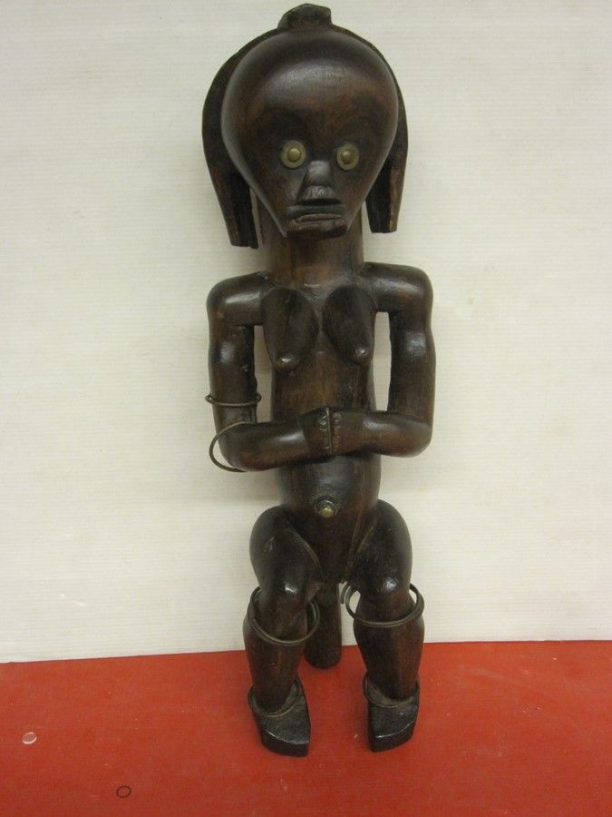 Figurine en bois sculpté, à grosse tête, bras croisés et anneaux métalliques aux membres.