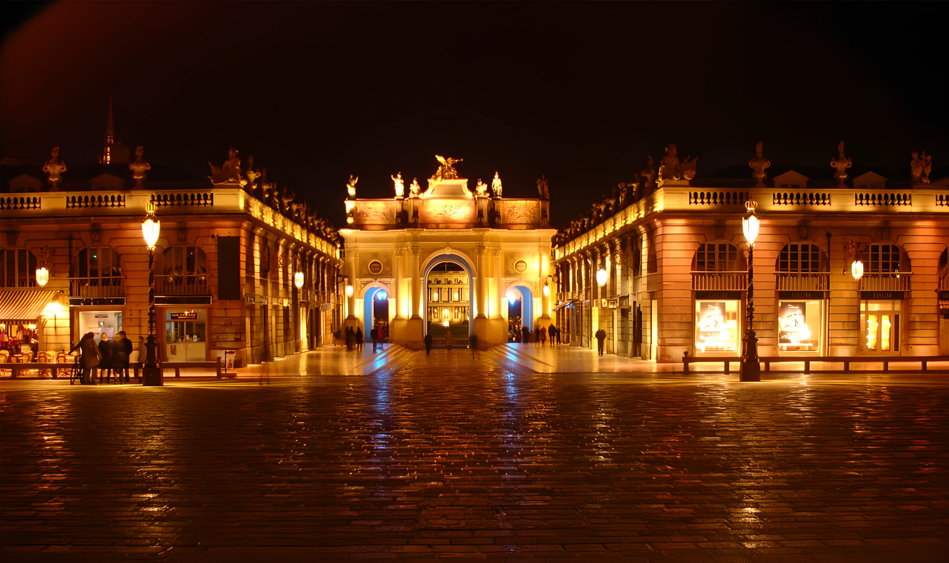Vue nocturne d'un grand bâtiment voûté flanqué de bâtiments symétriques, reflétant les lumières sur une surface humide.