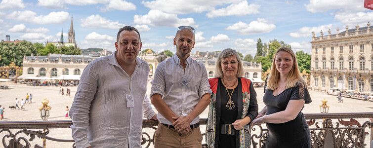 Quatre personnes se tiennent sur un balcon donnant sur une place avec un grand bâtiment et le clocher d'une église en arrière-plan.