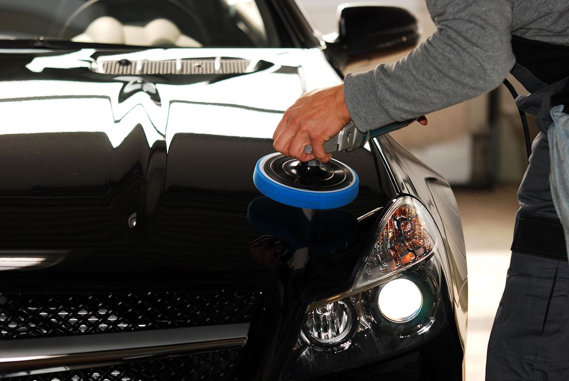 Dans un garage, une personne utilise une polisseuse électrique bleue pour lustrer le capot d'une voiture noire brillante.