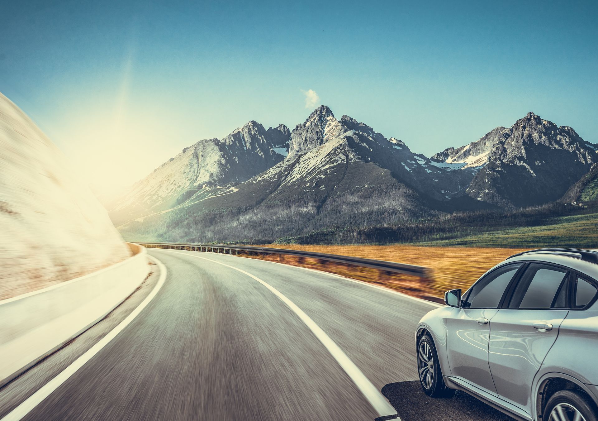 Une voiture argentée roule sur une route sinueuse en direction de montagnes enneigées et ensoleillées, sous un ciel d'un bleu limpide.
