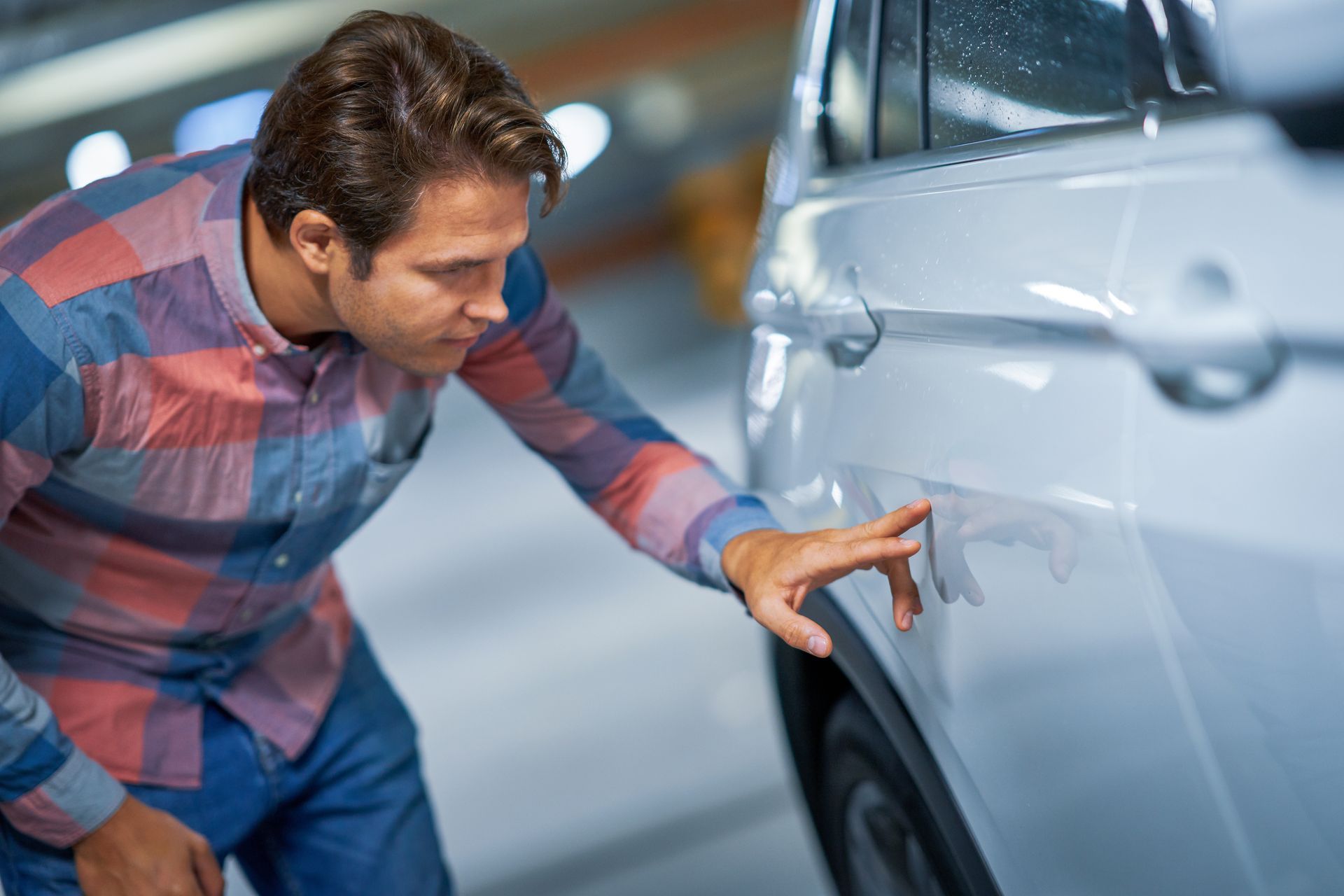Une personne portant une chemise à carreaux examine une bosse sur le côté d'un véhicule de couleur claire dans un parking souterrain.