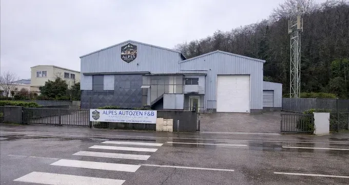 Un entrepôt industriel de couleur claire, orné d'un logo en forme de bouclier sur sa façade, situé derrière une entrée fermée et une route.