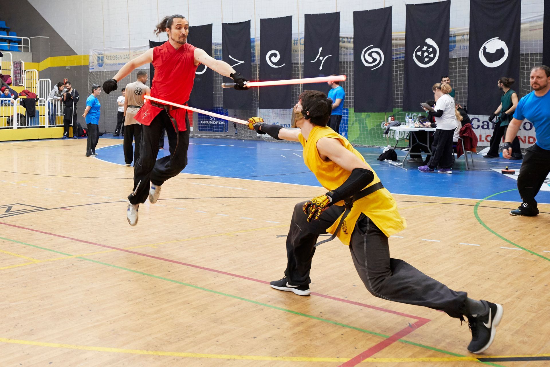 Dos personas se baten a duelo con armas acolchadas en un gimnasio. Uno con saltos rojos, el otro con estocadas amarillas.