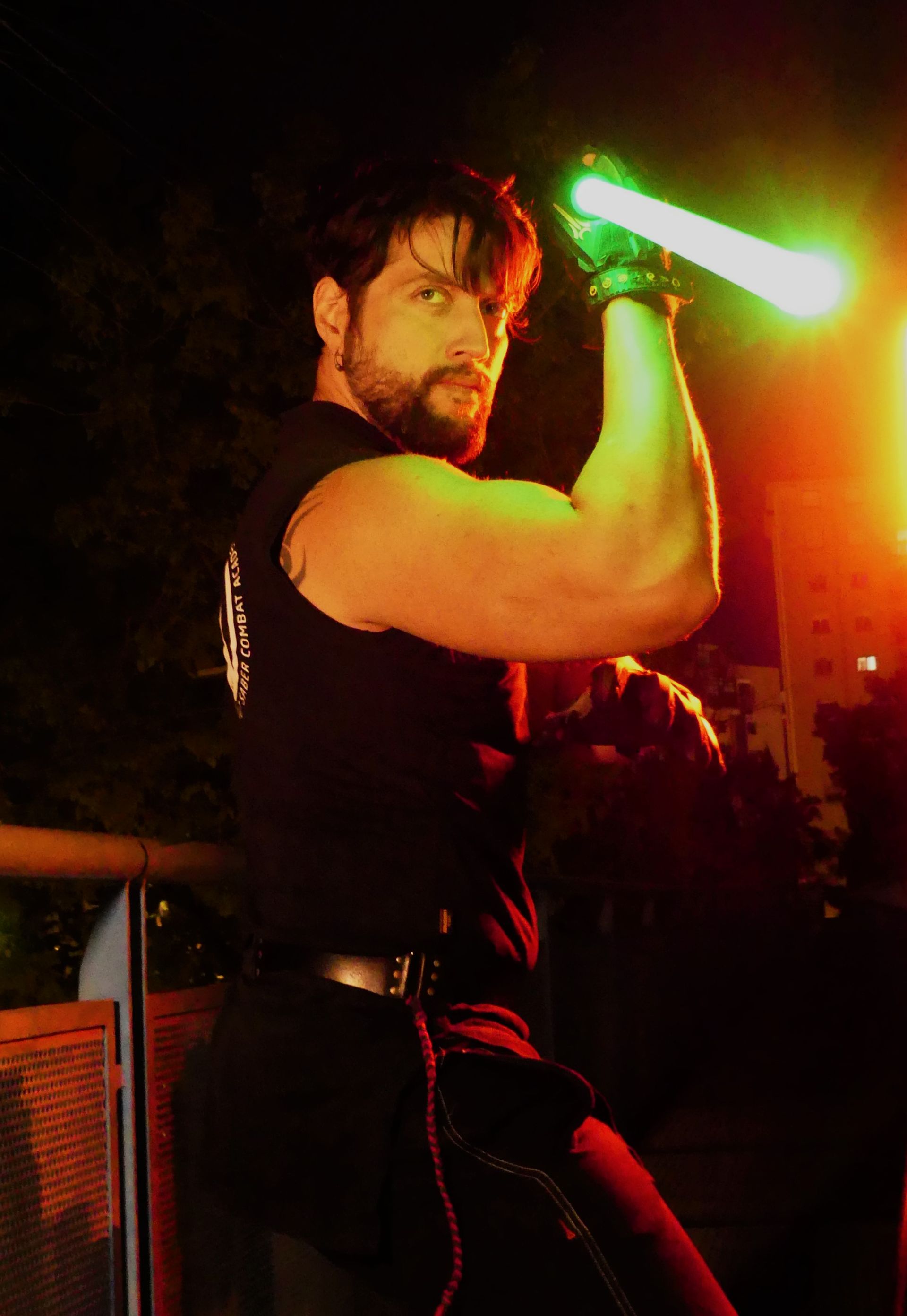 Un hombre con un sable de luz verde levanta su brazo musculoso; al aire libre, de noche.