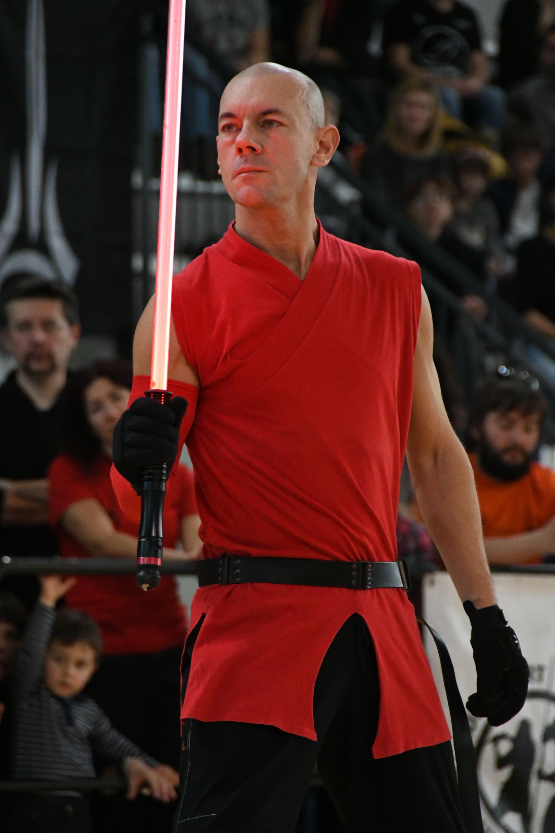 Hombre con camisa roja y guantes negros sosteniendo un sable de luz rojo, de pie entre una multitud.