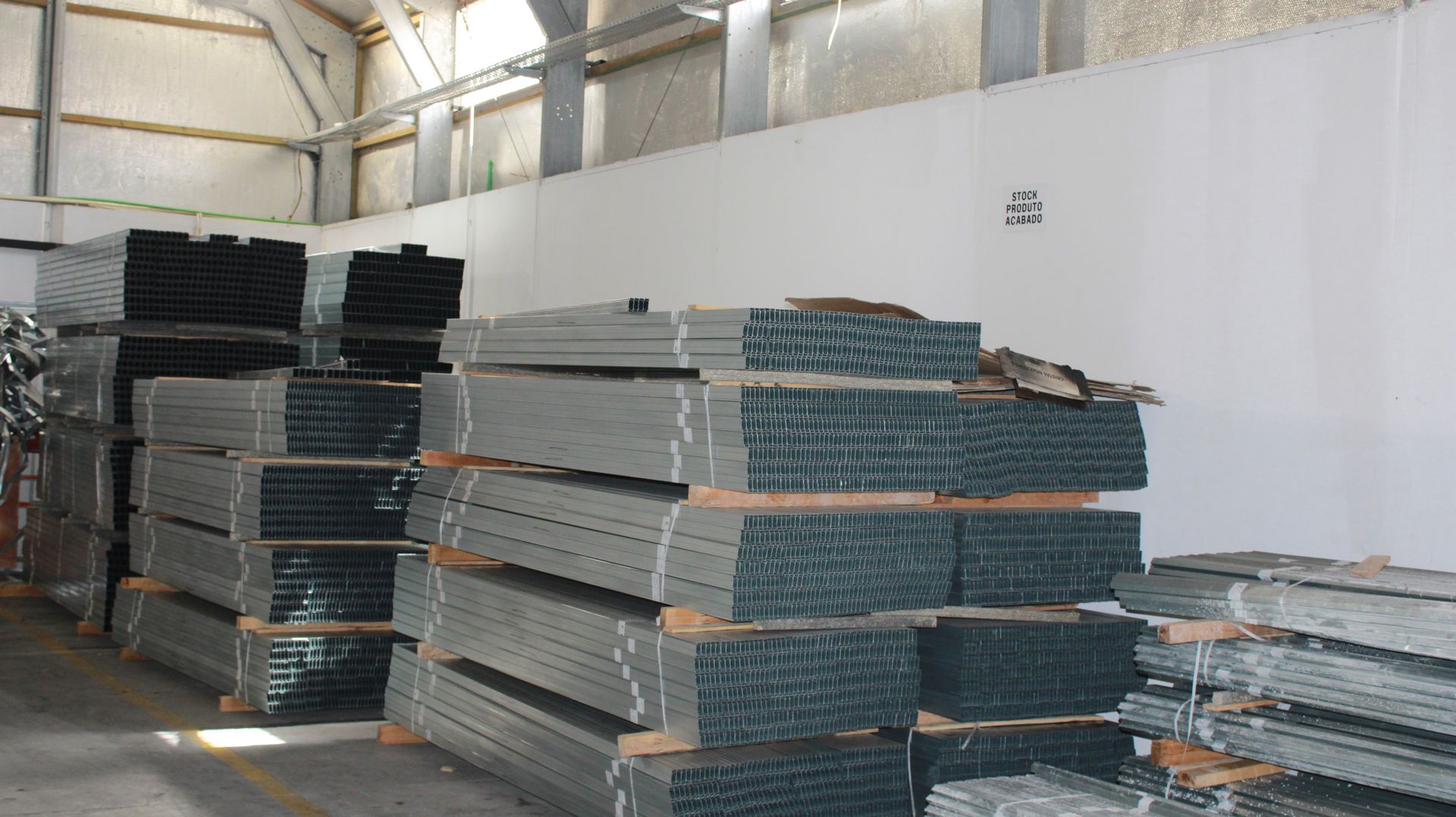 Pilhas de vigas de metal cinza armazenadas dentro de um armazém, com uma parede branca e um telhado de metal visíveis.