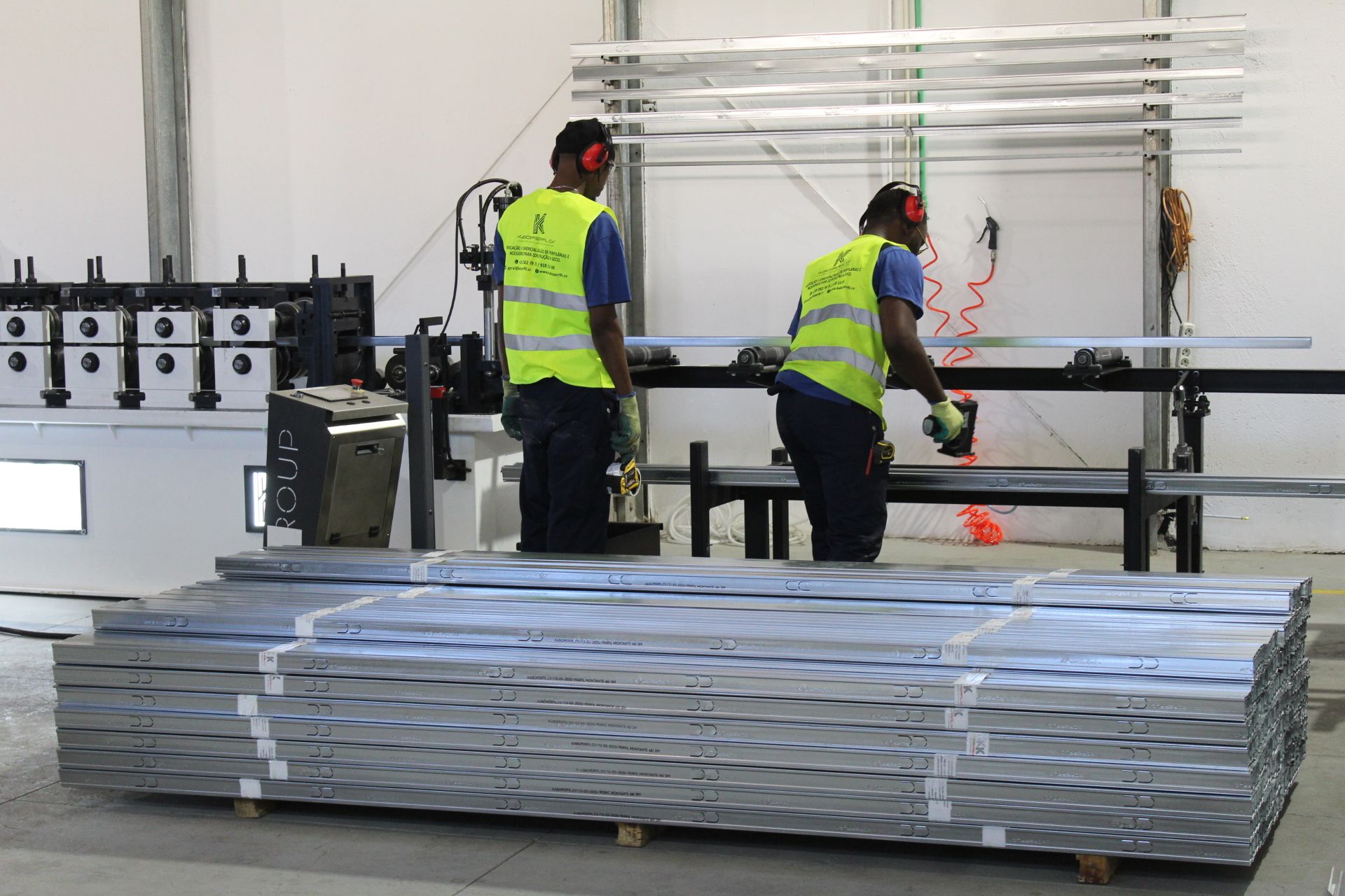 Trabalhadores com coletes de segurança usam ferramentas em chapas de metal em uma fábrica.