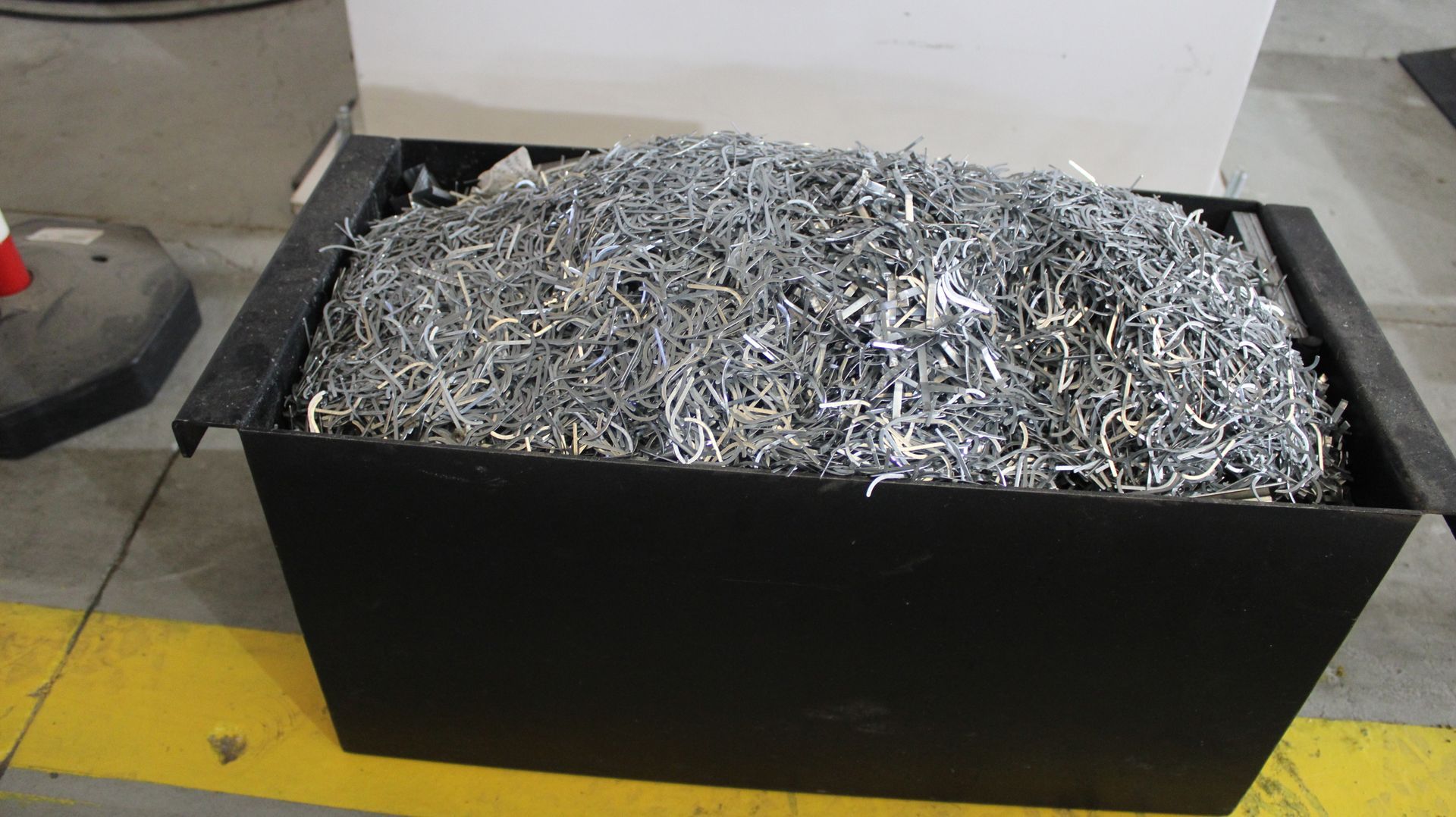 Lixeira preta transbordando de aparas de metal, no chão de uma fábrica.