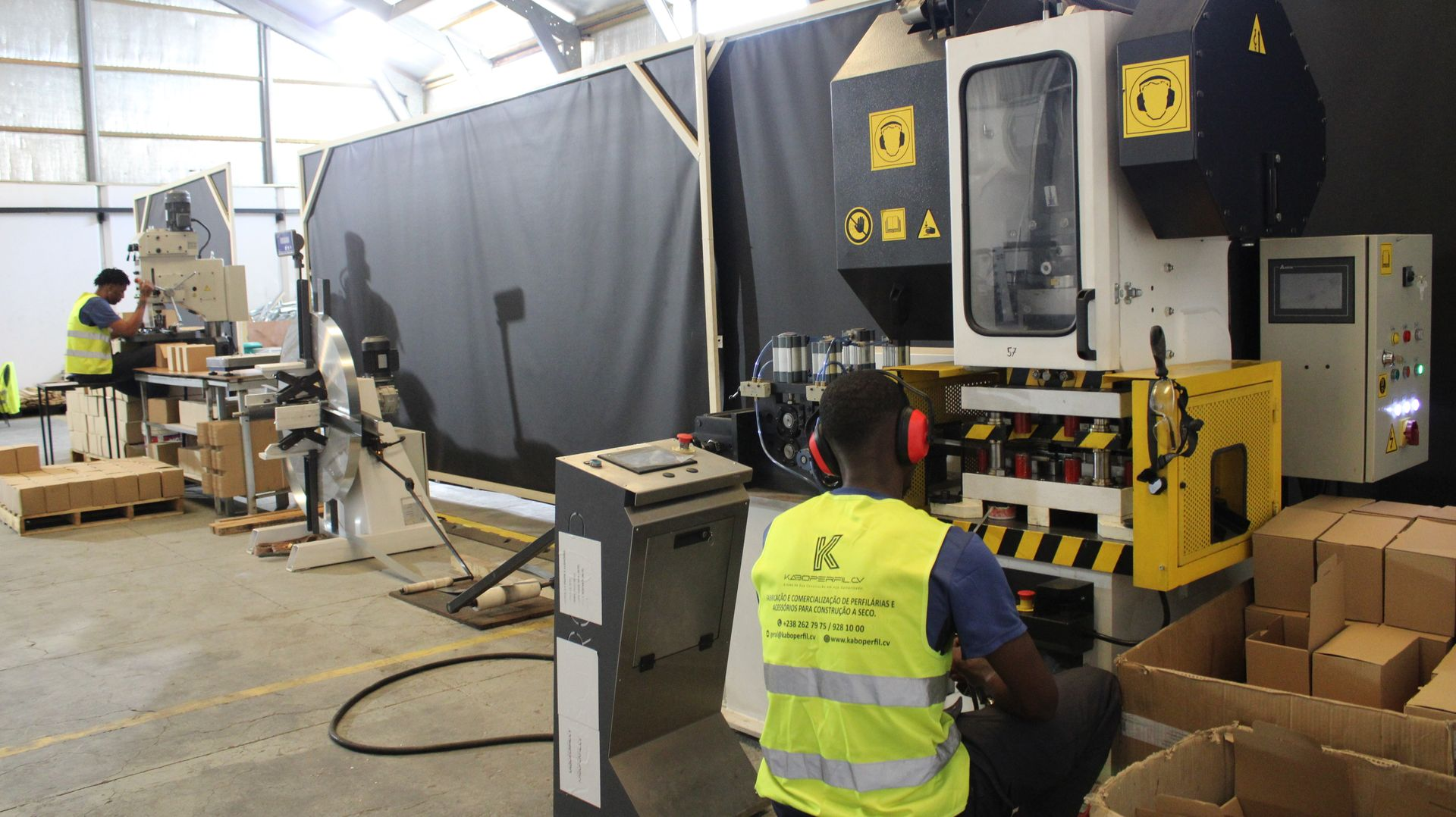 Trabalhadores operando máquinas industriais em uma fábrica. Um trabalhador usa um colete de segurança amarelo.