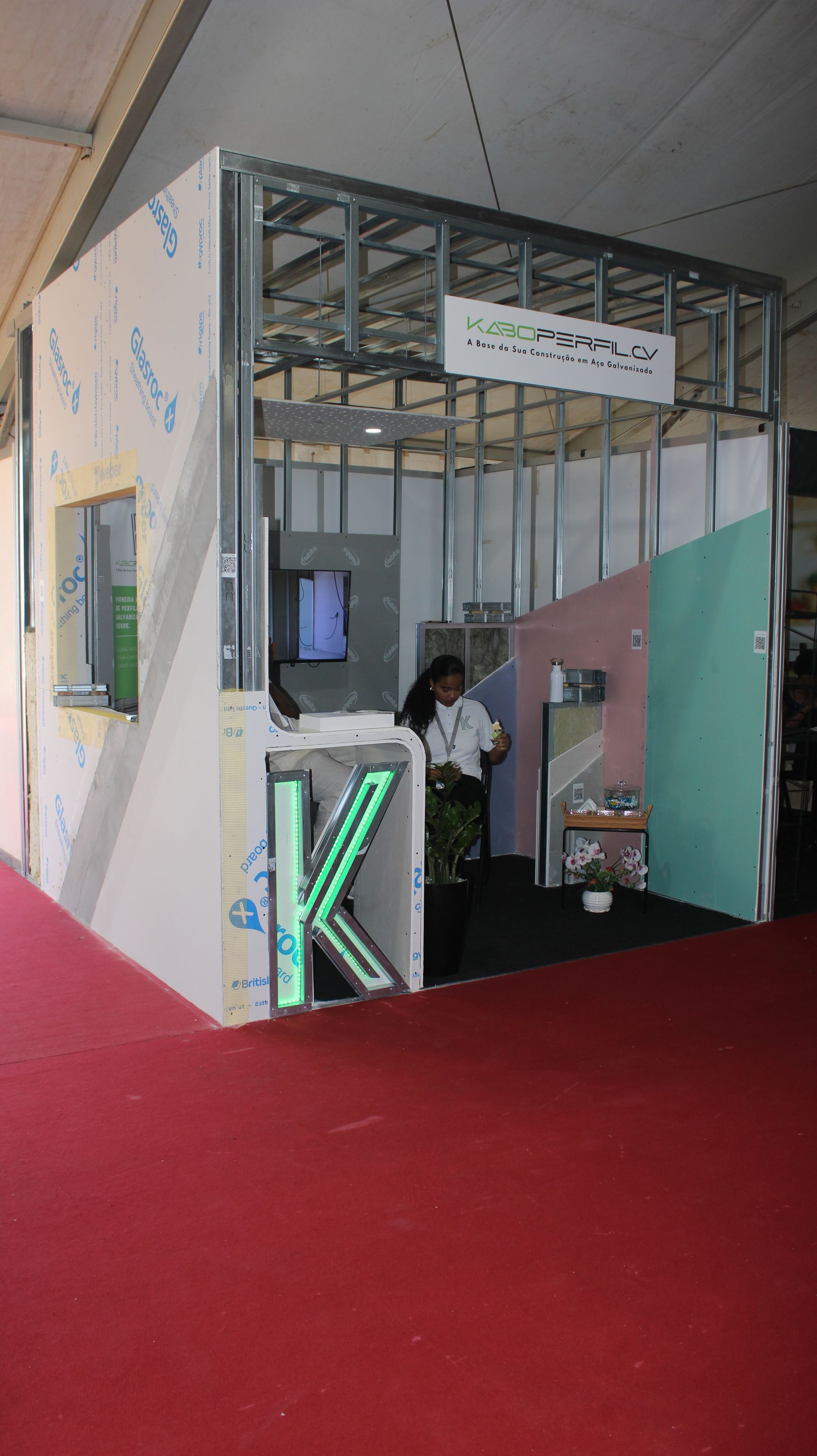 Cabine com estrutura metálica exposta, paredes parcialmente construídas, carpete vermelho e uma pessoa dentro.