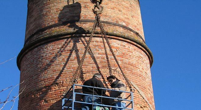 Dos personas en una cesta de construcción azul suspendida por una grúa, reparando una alta chimenea de ladrillos contra un cielo azul.