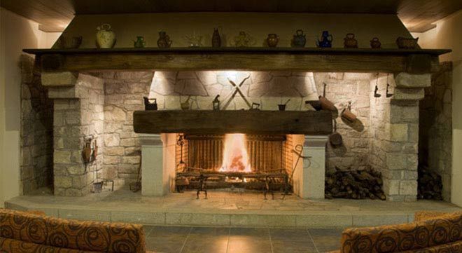 Una gran chimenea rústica de piedra con un fuego resplandeciente, coronada por una repisa de madera con elementos decorativos y herramientas en forma de cruz.