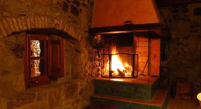 Una habitación de piedra con una chimenea encendida y una ventana de madera arqueada con contraventanas abiertas.