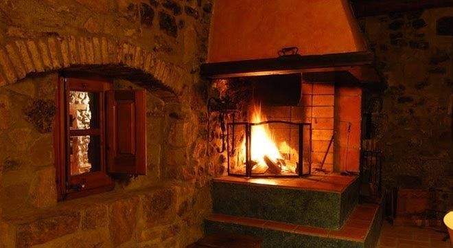 Una habitación cálida con paredes de piedra cuenta con una chimenea con un fuego brillante detrás de una mampara junto a una ventana abierta y empotrada.