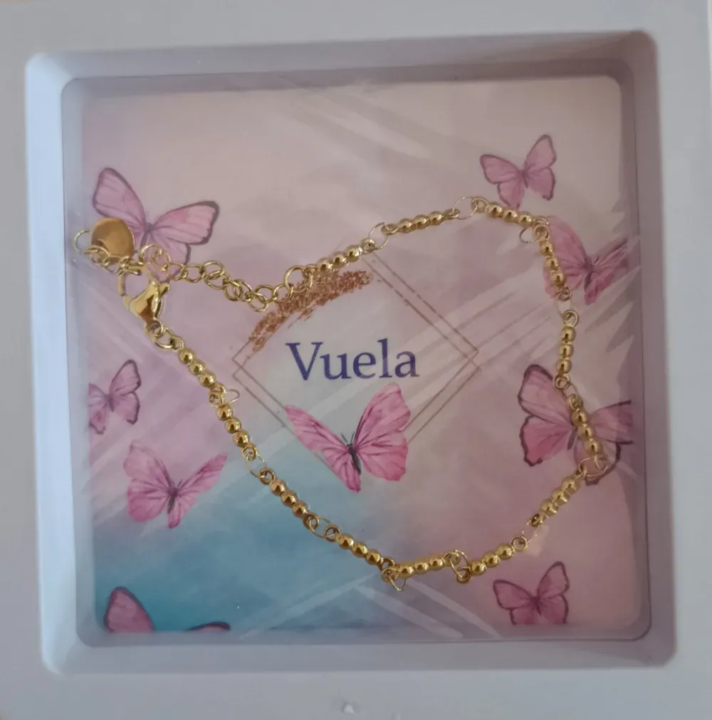 Pulsera dorada con una mariposa sobre una tarjeta en tonos pastel con motivos de mariposas y la palabra «Vuela» centrada.
