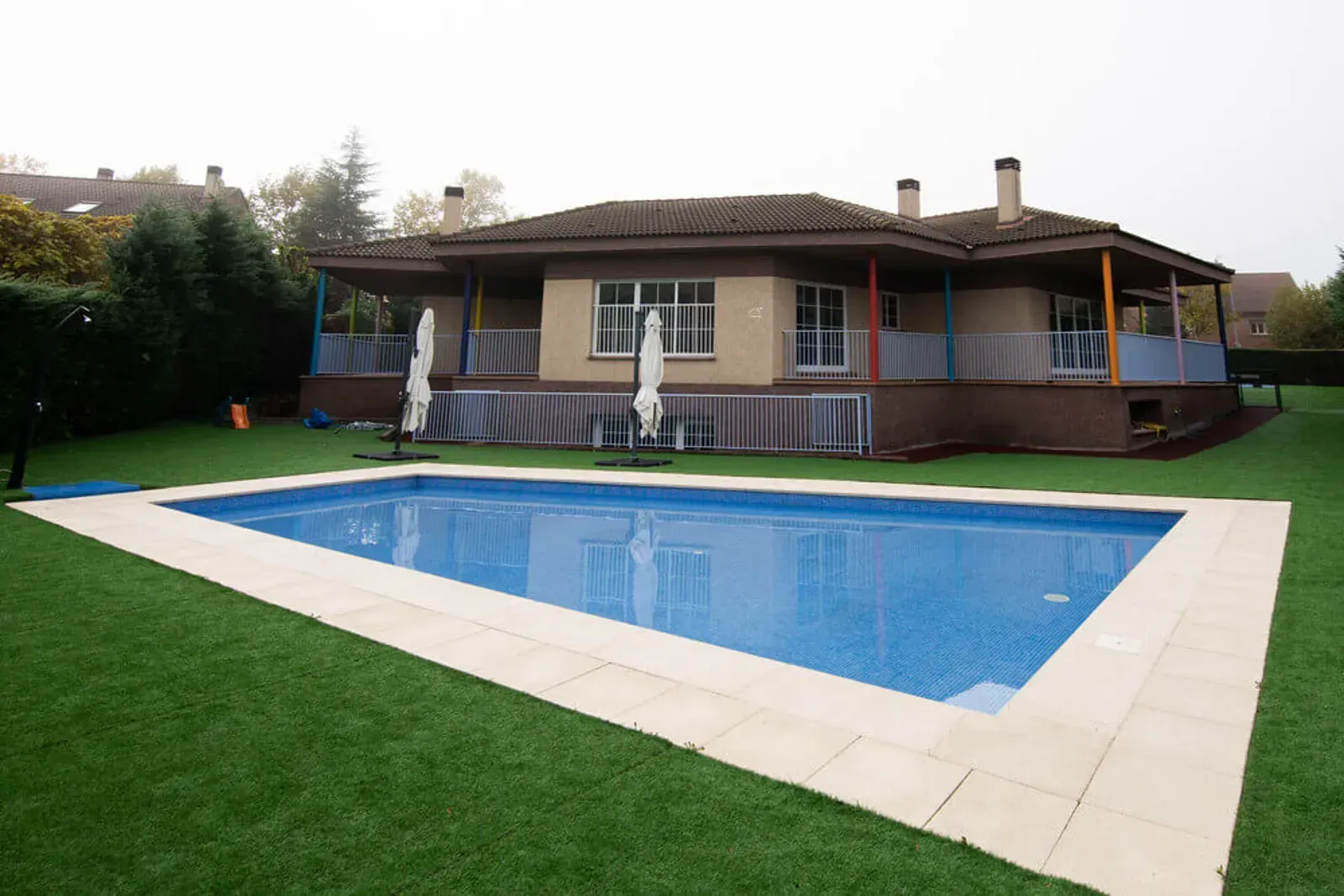 Una casa con porche y piscina. La piscina tiene azulejos azules y la casa tiene el techo marrón.